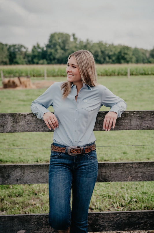A woman stands outdoors leaning against a wooden fence in a grassy field under an overcast sky. She is wearing a light blue long-sleeve button-up shirt, dark blue jeans, and a brown belt.