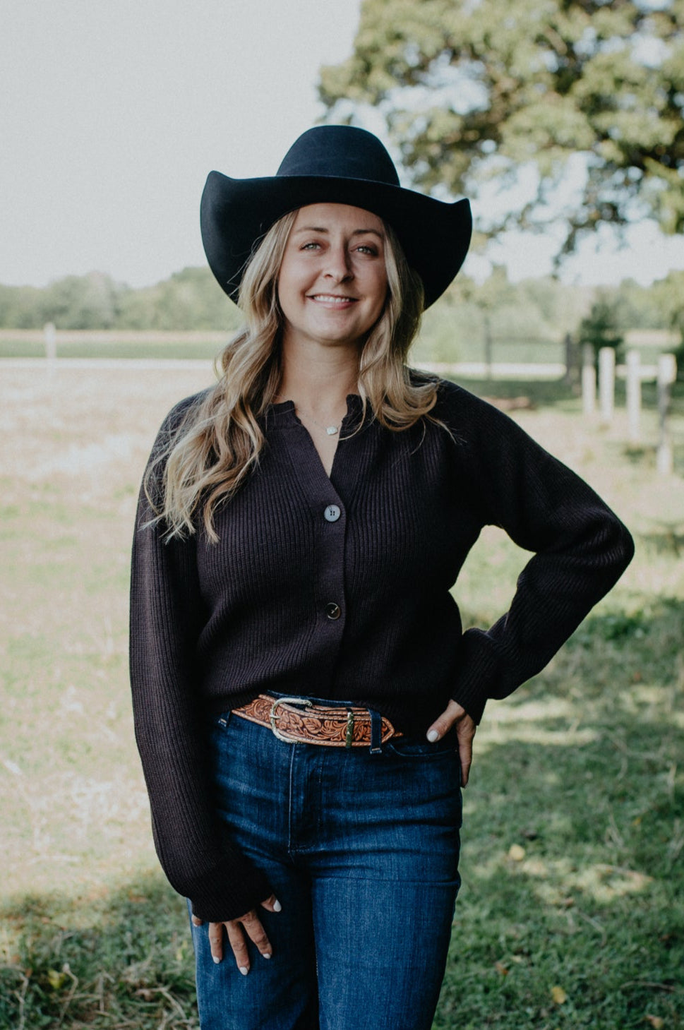 Woman wearing a black cowboy hat, black cardigan, and blue jeans standing in a grassy field.