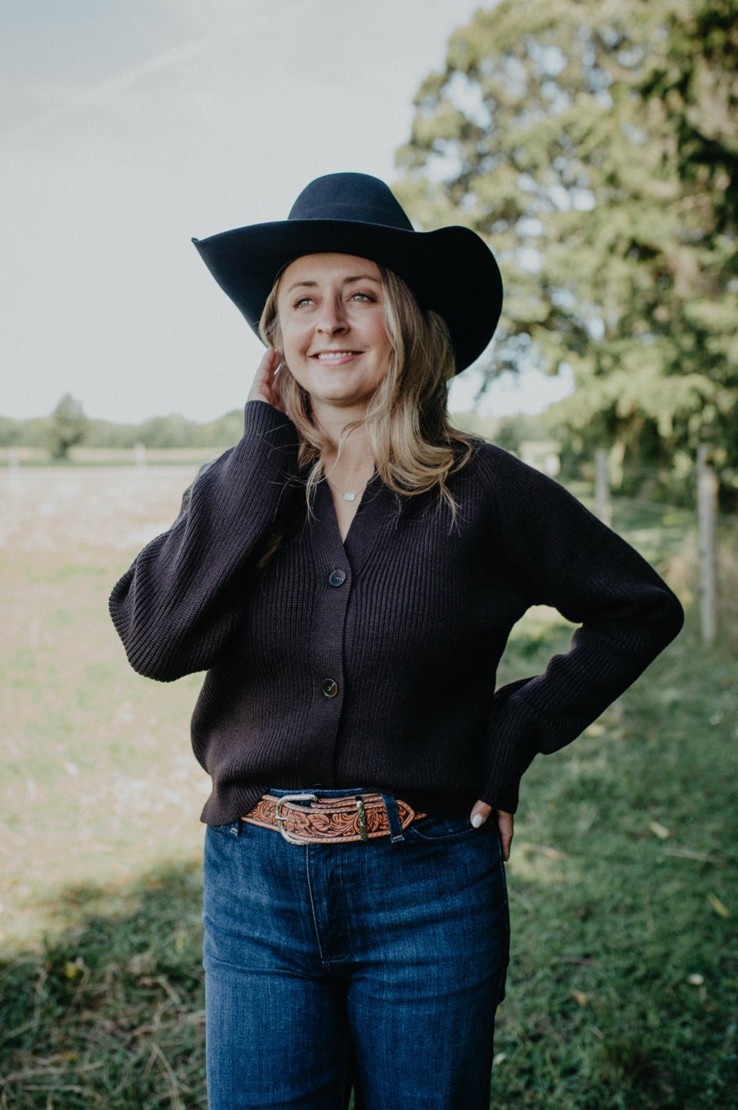 Woman wearing a black cowboy hat, dark shirt, and blue jeans standing in a field with trees in the background.